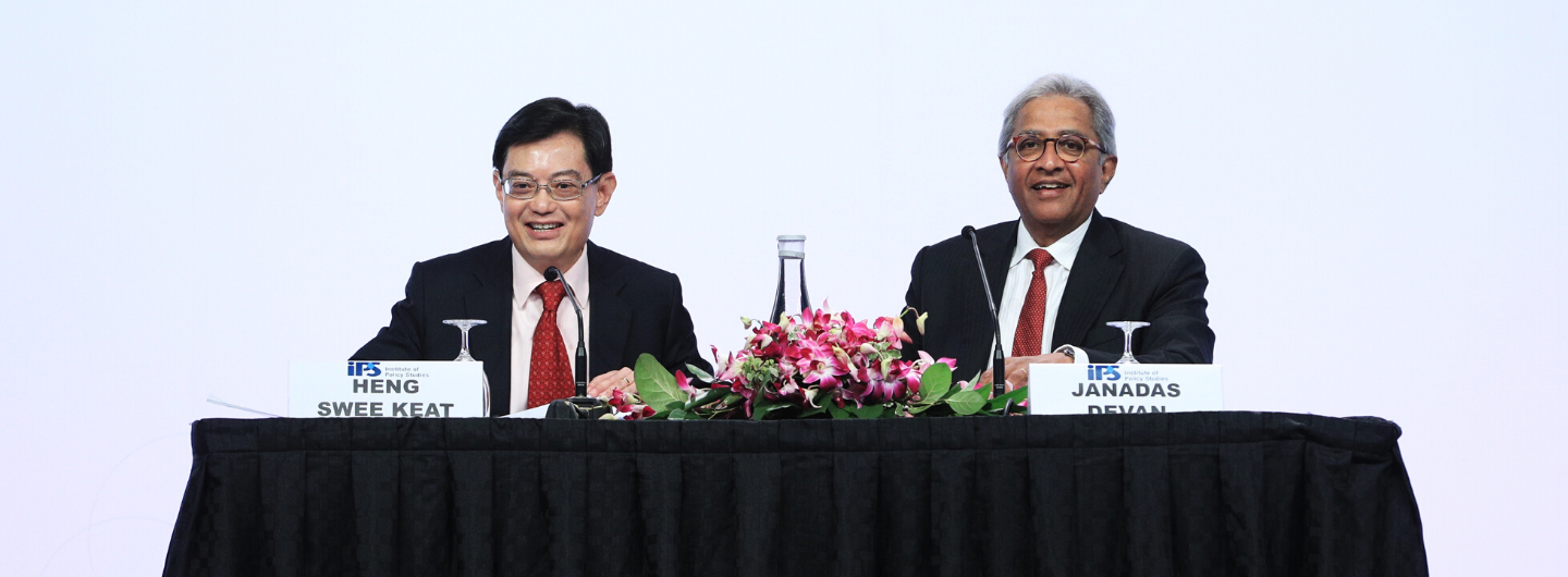 Two men in suits seated at table with microphones. "Heng Swee Keat" and "Janadas Devan" nameplates.